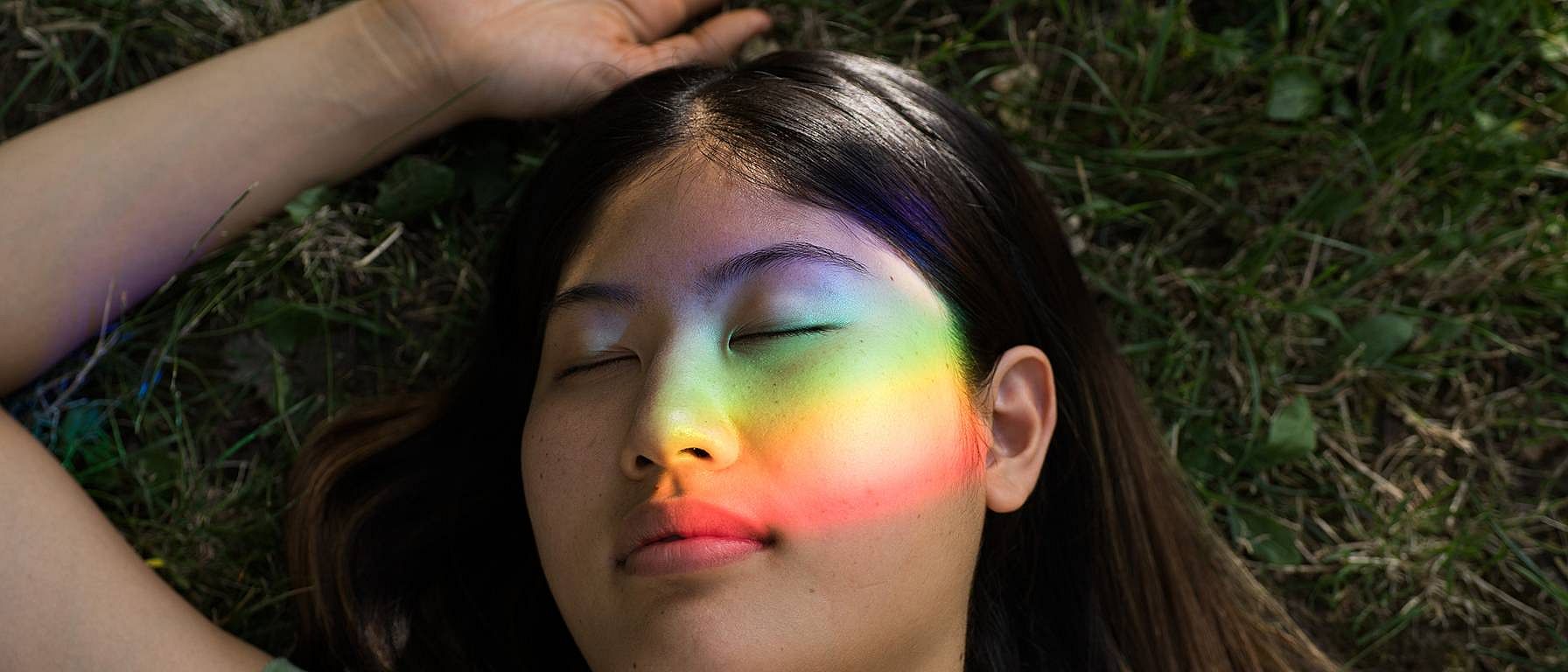 Frau liegt im Gras und ein Regenbogenlicht leuchtet über das Gesicht.