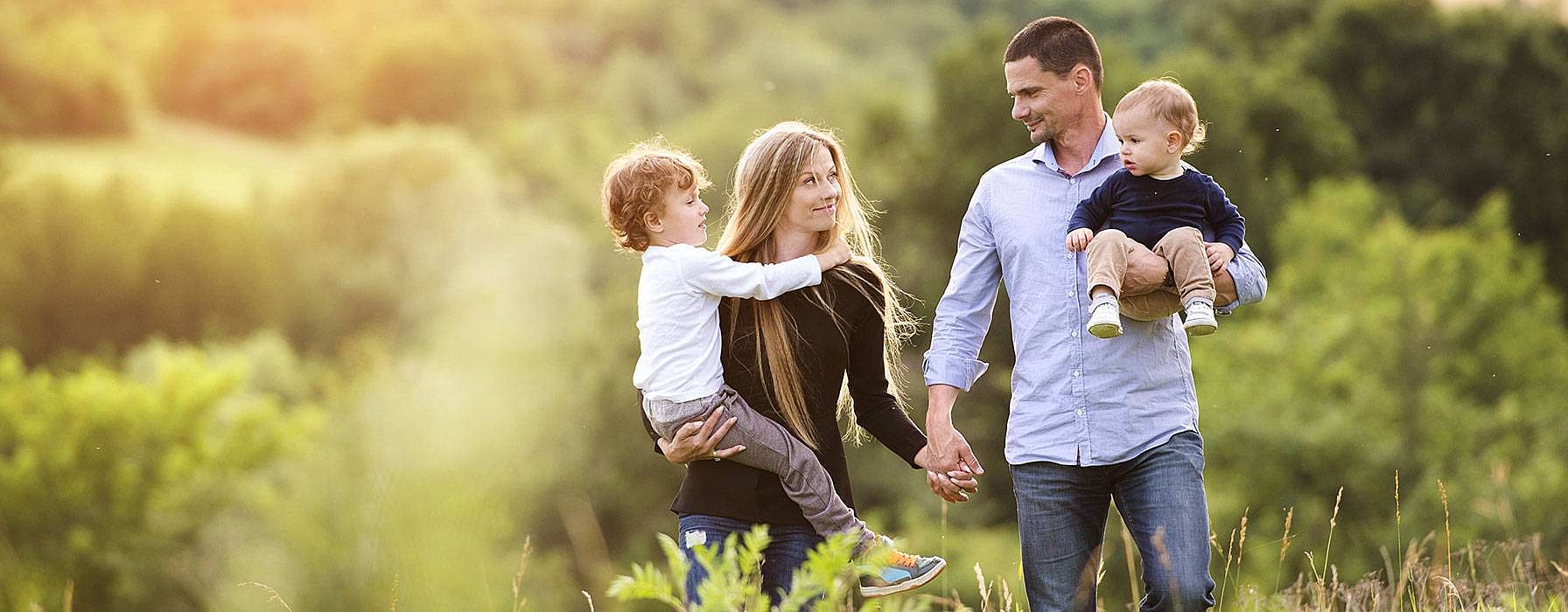 Mann und Frau spazieren Händchenhaltend mit jeweils einem Kind auf dem Arm in der Natur
