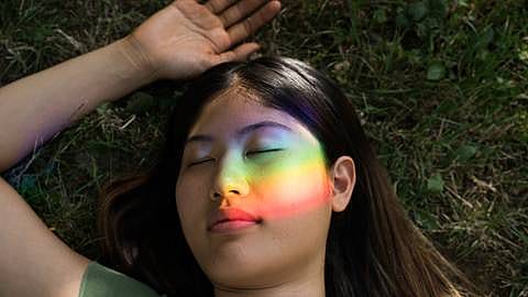 Frau liegt im Gras und ein Regenbogenlicht leuchtet über das Gesicht.
