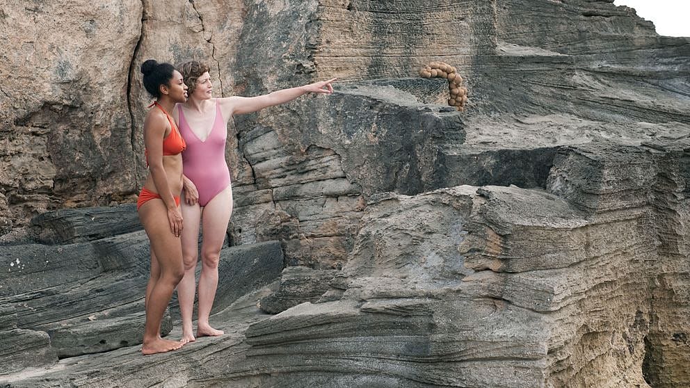 Zwei Frauen in Badekleidung stehen am Felsen und schauen in die Ferne